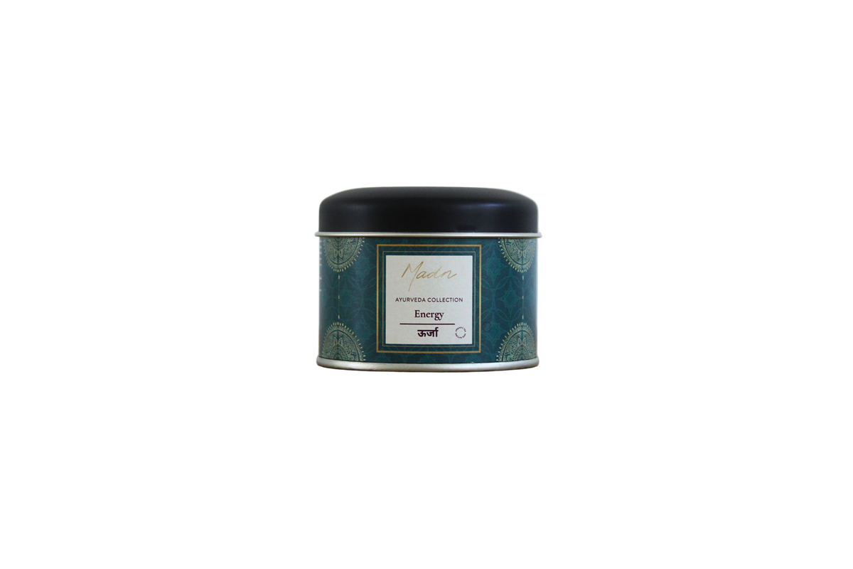 A small round tin candle labeled 'Maison Nicole Energy' sits on a light wooden surface against a plain light gray background.